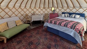 A yurt interior with a round ceiling, a bed with a patterned blanket, a small table with a checkered tablecloth, and a chair. - Dragon Orchard Retreat (Ledbury)