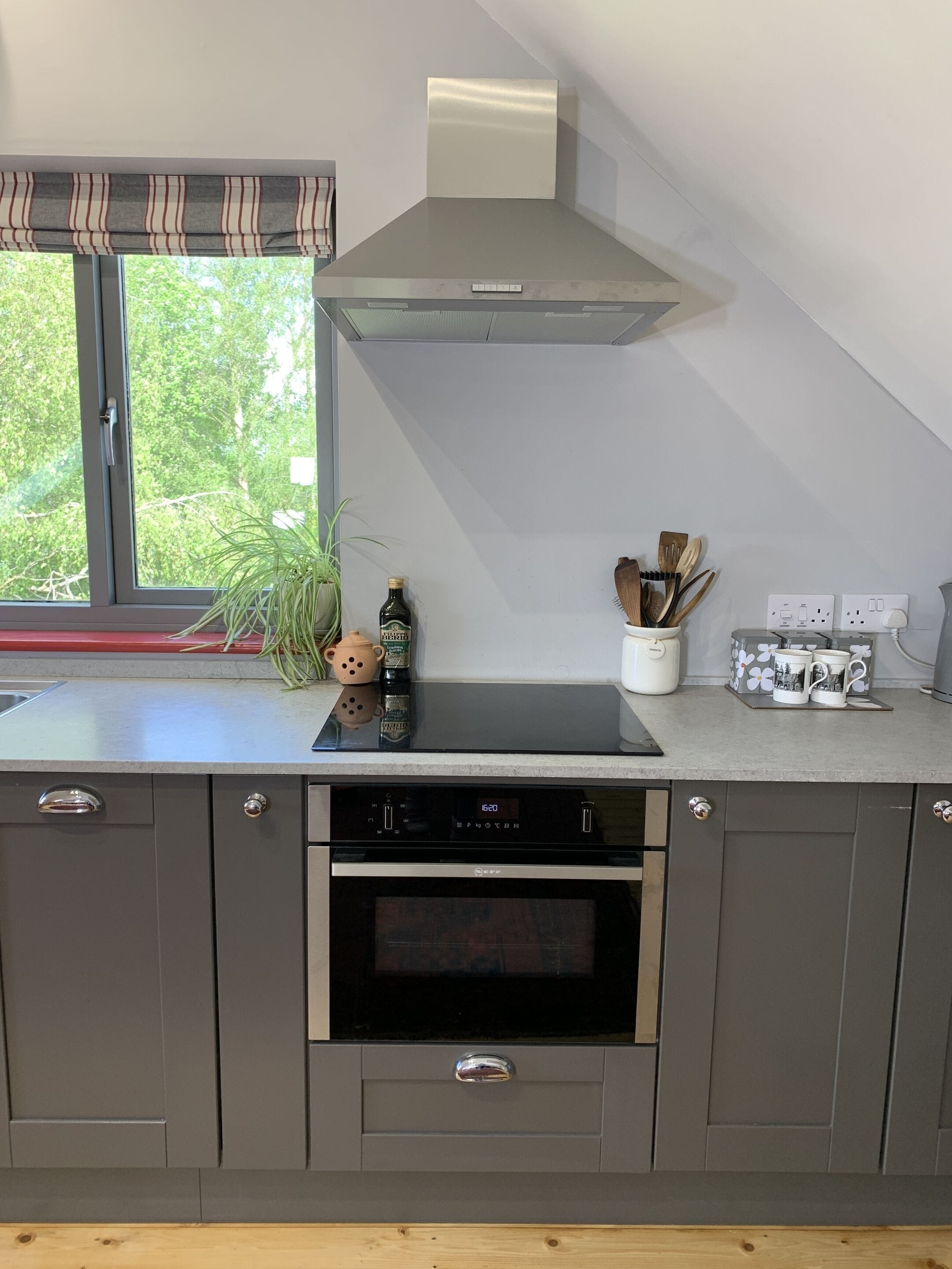 A modern kitchen with grey cabinets, a built-in oven, a stainless steel range hood, and a window with a view of greenery.