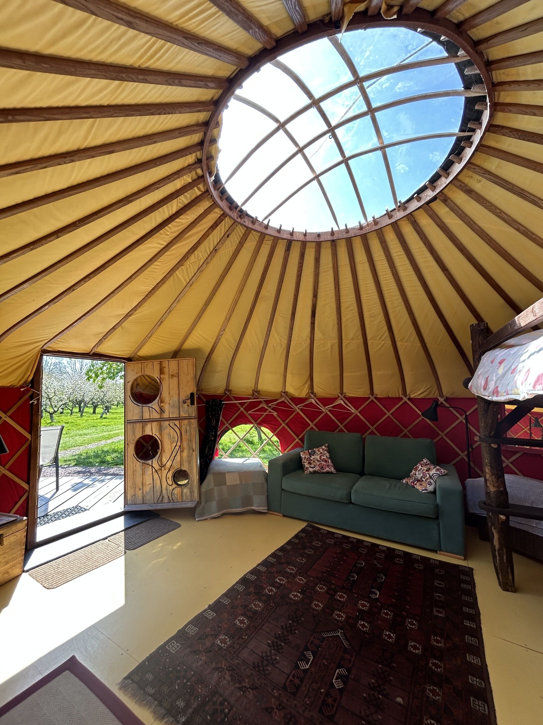 Interno di una yurta con un lucernario centrale, un divano verde e un tappeto decorato.