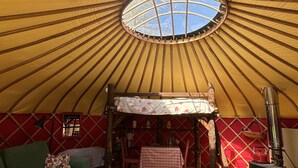 A yurt interior with a round ceiling, a central sleeping area with a bed, a small table, a sofa, a rug, and a wood-burning stove. - Dragon Orchard Retreat (Ledbury)