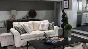A modern living room with a white sofa, patterned cushions, a coffee table with decorative items, a large abstract painting, and a chandelier.
