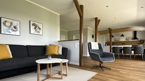Un salon moderne avec un canapé gris foncé, une petite table blanche et un fauteuil gris. Le plafond est orné de poutres en bois et, au fond, on aperçoit un coin cuisine.