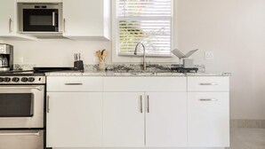 A modern kitchen with white cabinets, a built-in oven, a microwave, and a window with blinds. - Paradise on the Bay  (Goodland)