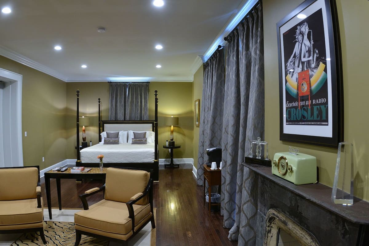 A hotel room with a four-poster bed, a sitting area with two chairs, a small table, a fireplace, and a framed poster on the wall.