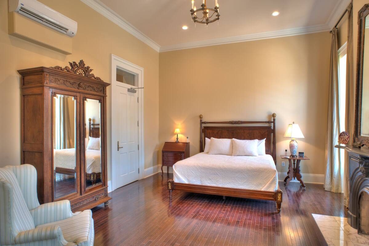 A spacious bedroom with a large bed, a wooden wardrobe, a sitting area with a chair, and a fireplace.