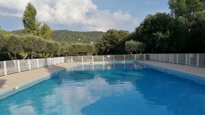 A rectangular swimming pool with a white fence surrounding it, set against a backdrop of trees and hills.