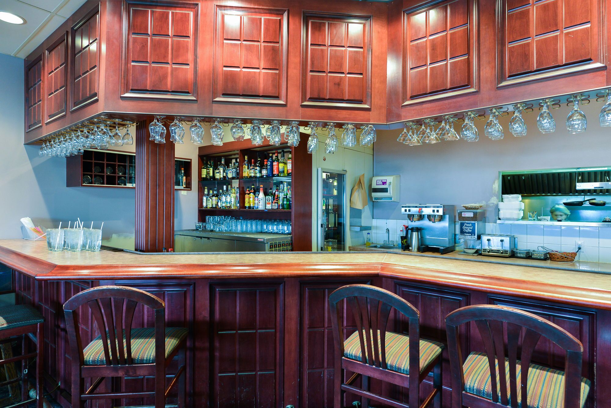 A bar area with wooden cabinets, a counter, and bar stools. There are shelves with bottles and glasses, and a coffee machine.