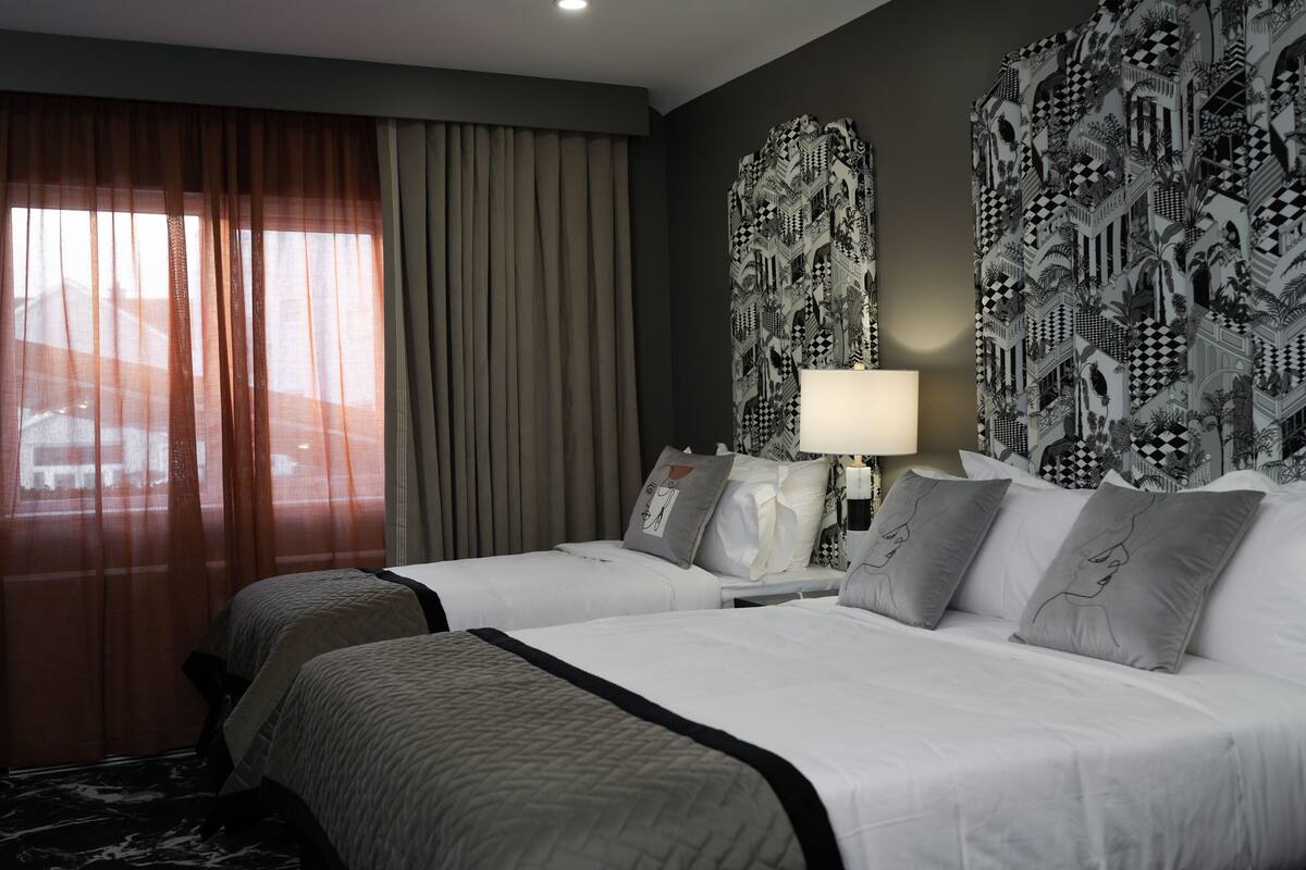 A hotel room with two beds, a large window with sheer curtains, a patterned wall, and a nightstand with a lamp.