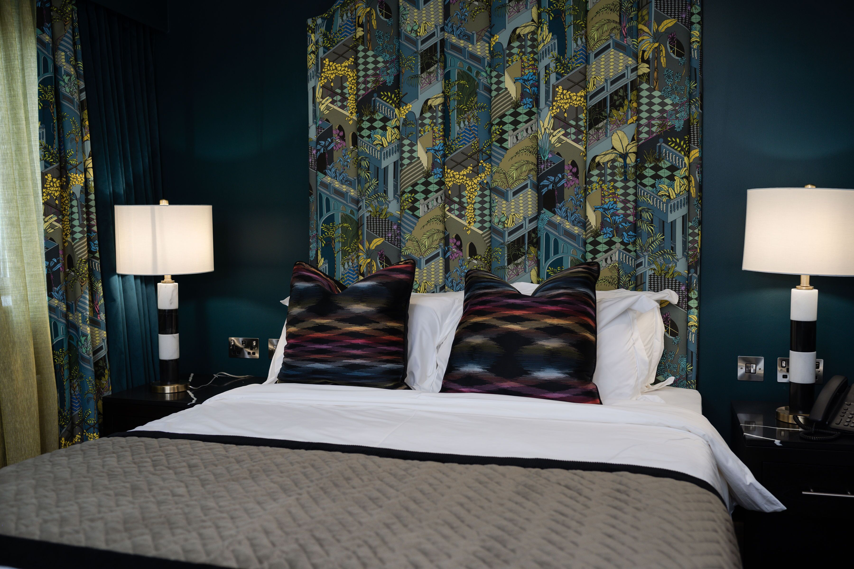 Quarto de hotel com uma cama grande, duas mesas de cabeceira com abajures, e uma parede estampada atrás da cama.
