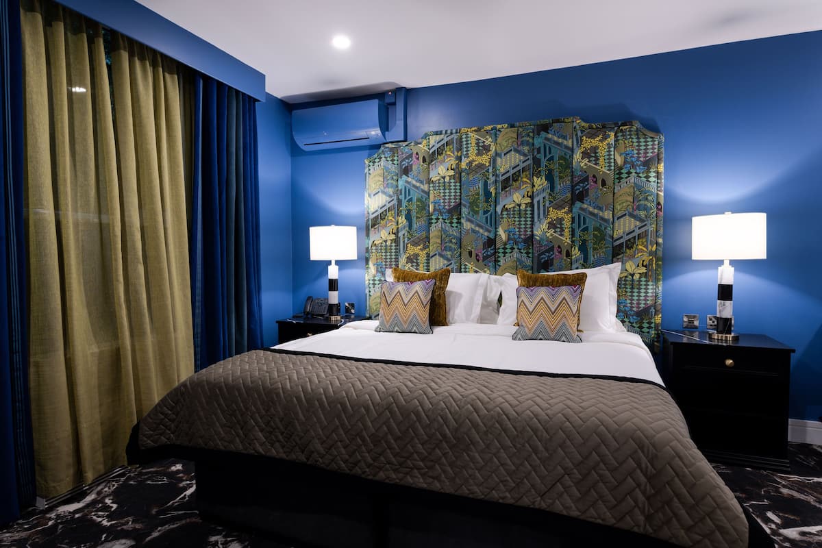 A hotel room with a large bed, two bedside tables, a headboard with a colorful pattern, and a window with curtains.