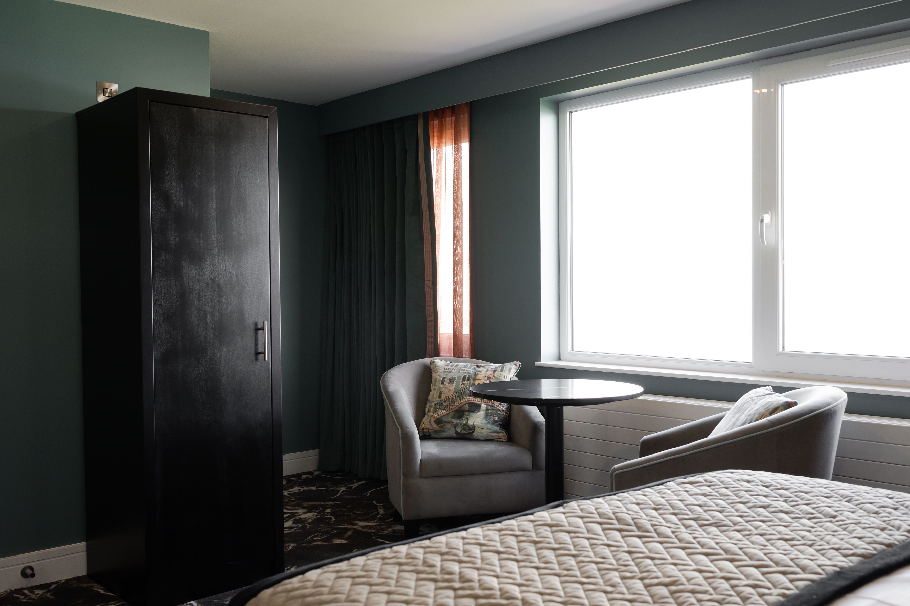 A hotel room with a bed, two armchairs, a small table, a wardrobe, and a window with curtains.