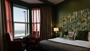 A hotel room with a large bed, a chair, a small table, a lamp, and a view of the ocean through a window with curtains. - Elephant Rock Hotel (Portrush)