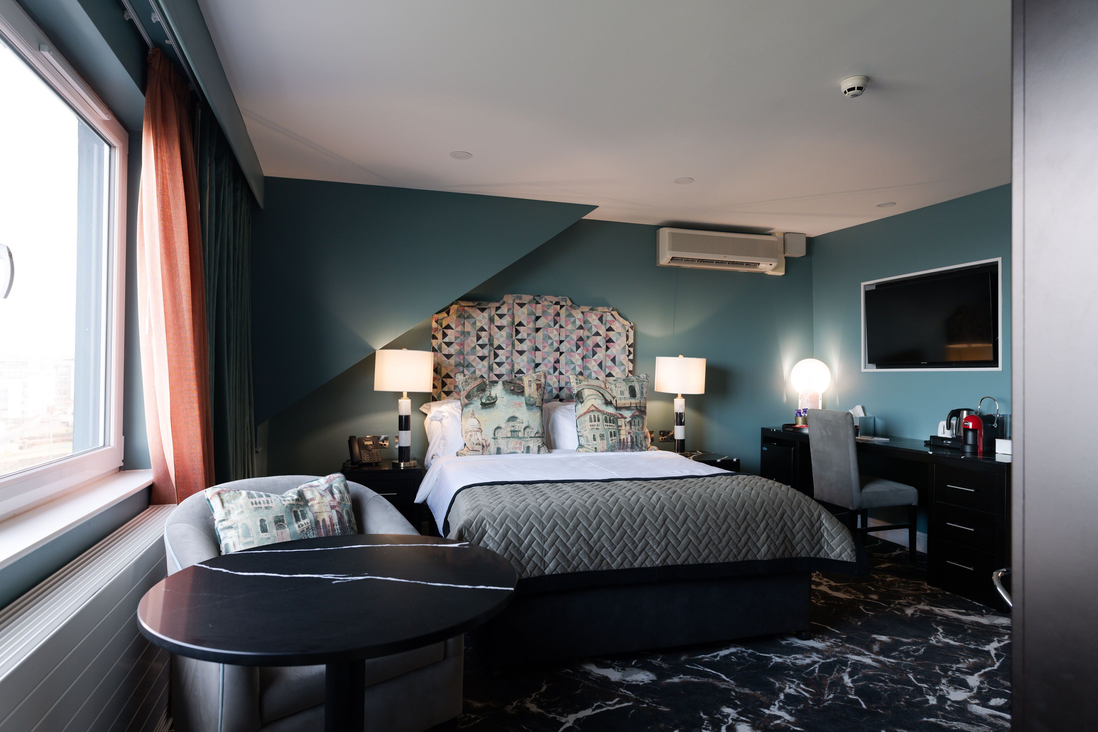 Quarto de hotel com cama, duas luminárias de cabeceira, escrivaninha com cadeira, mesinha e uma janela grande com cortinas.