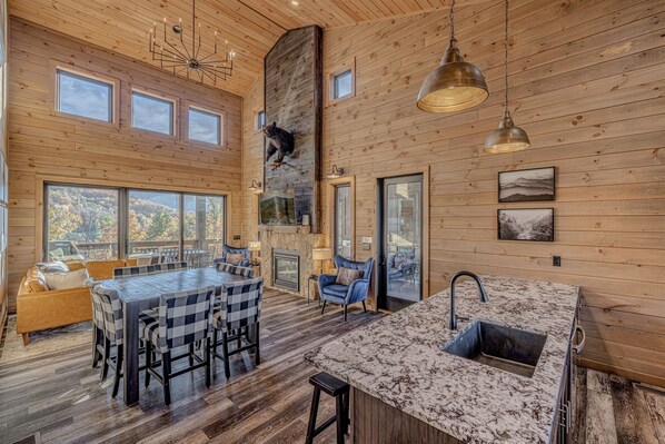 Un intérieur spacieux en bois, avec une grande table à manger, un îlot de cuisine doté d’un évier et une cheminée.