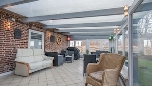 Een overdekt terras met een bakstenen muur, een witte bank, rieten stoelen en een glazen deur die naar buiten leidt.