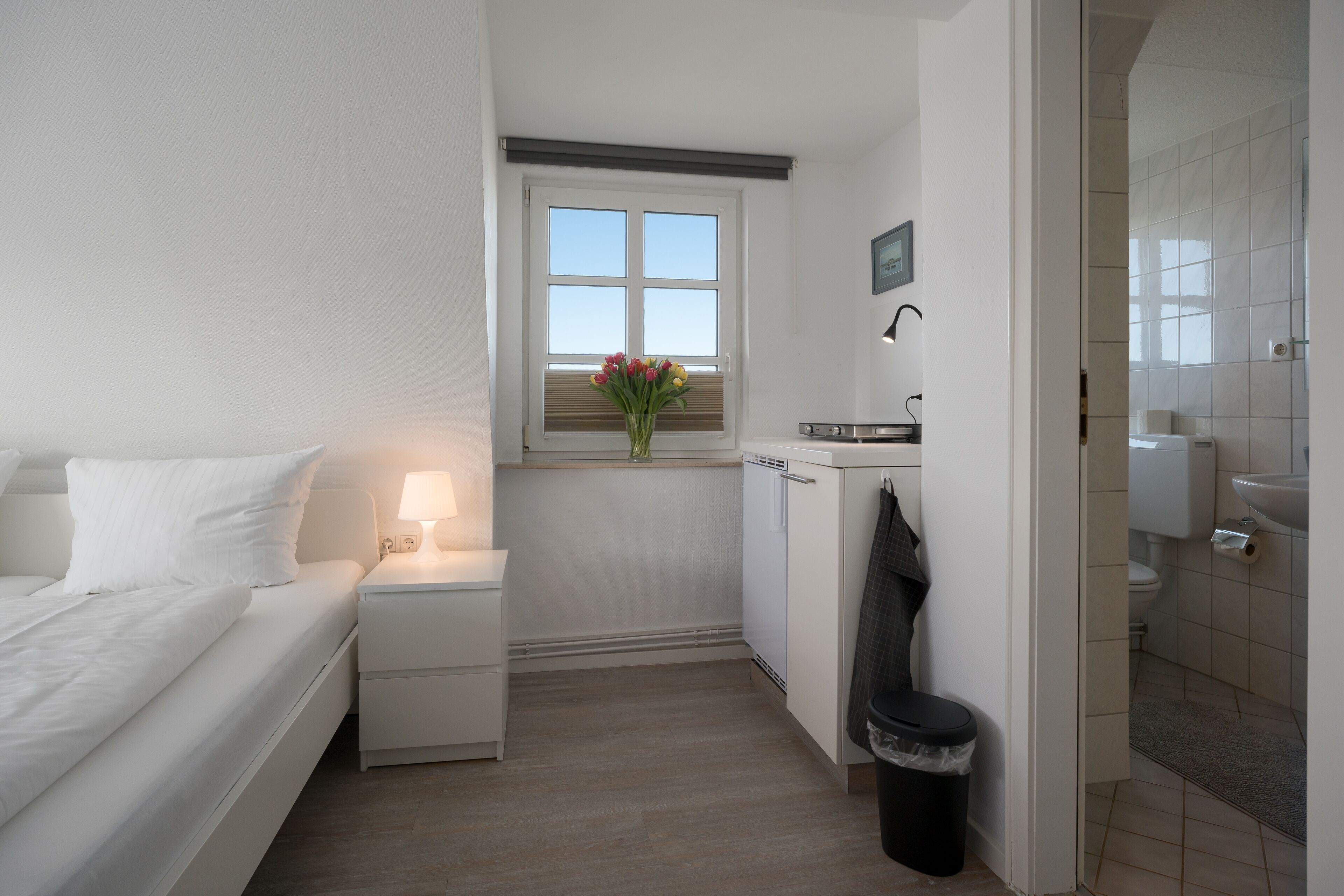 A small, compact bedroom with a bed, bedside table, lamp, and a window with a view of the sky.