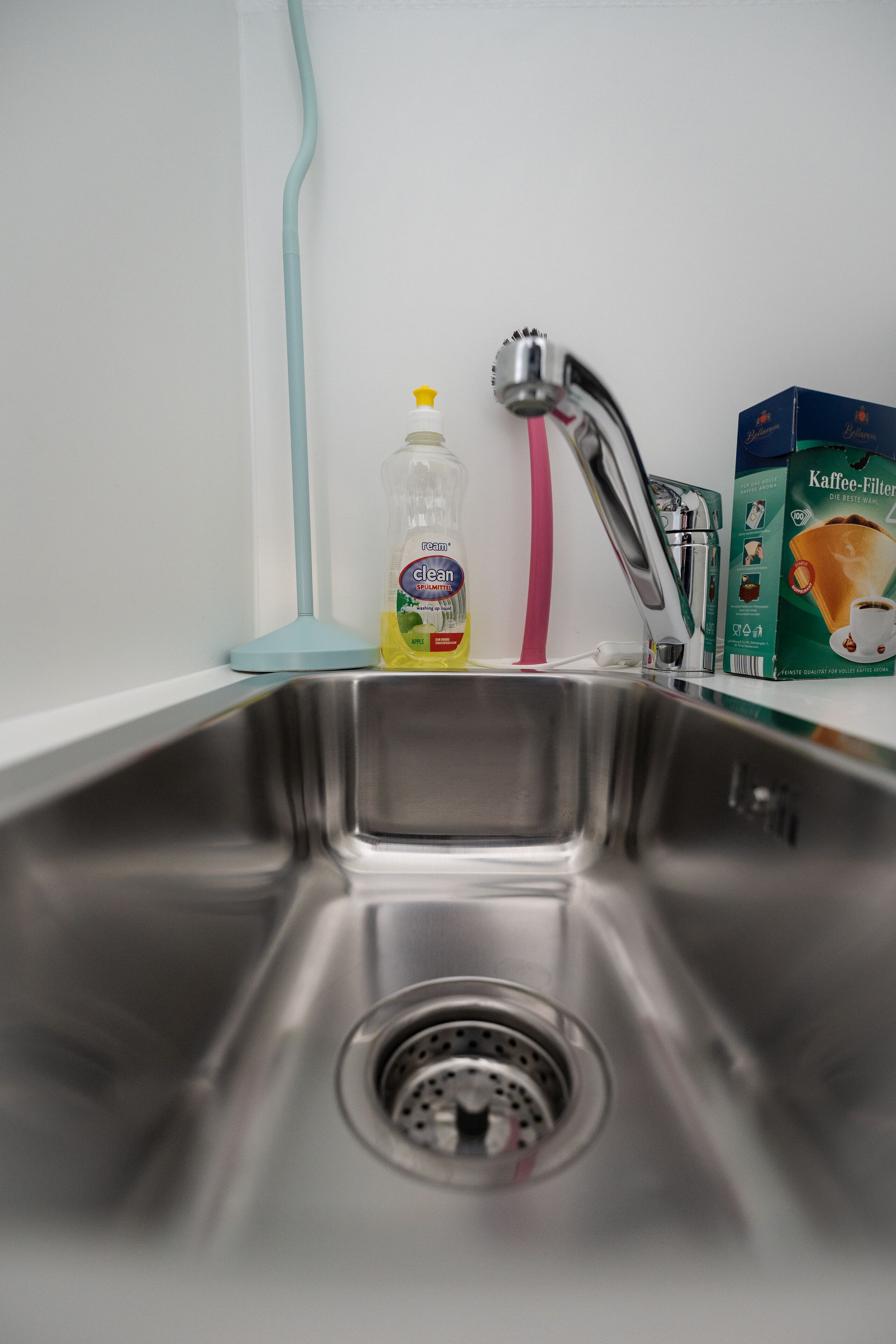 Un lavamanos de acero inoxidable con desagüe, una botella de detergente, un cepillo de limpieza rosa y una caja de filtros de café.