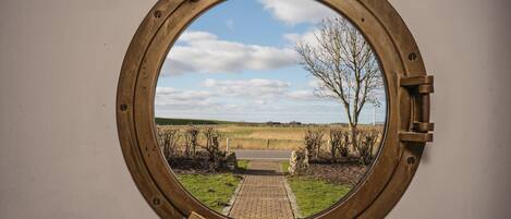 A porthole window with a view of a rural landscape, including a pathway and trees.
