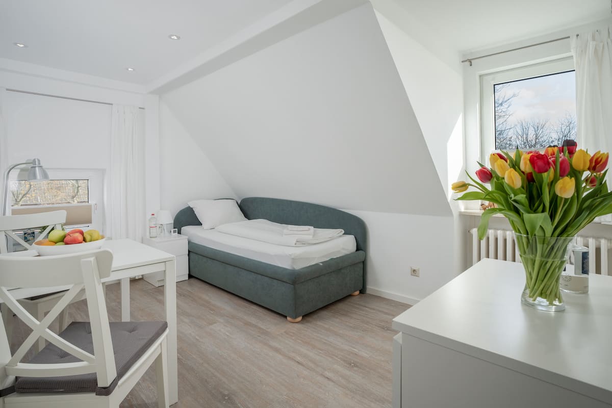 Een moderne slaapkamer met een bed, een bureau en een stoel. Er is een raam met uitzicht op bomen en een vaas met bloemen op het bureau.