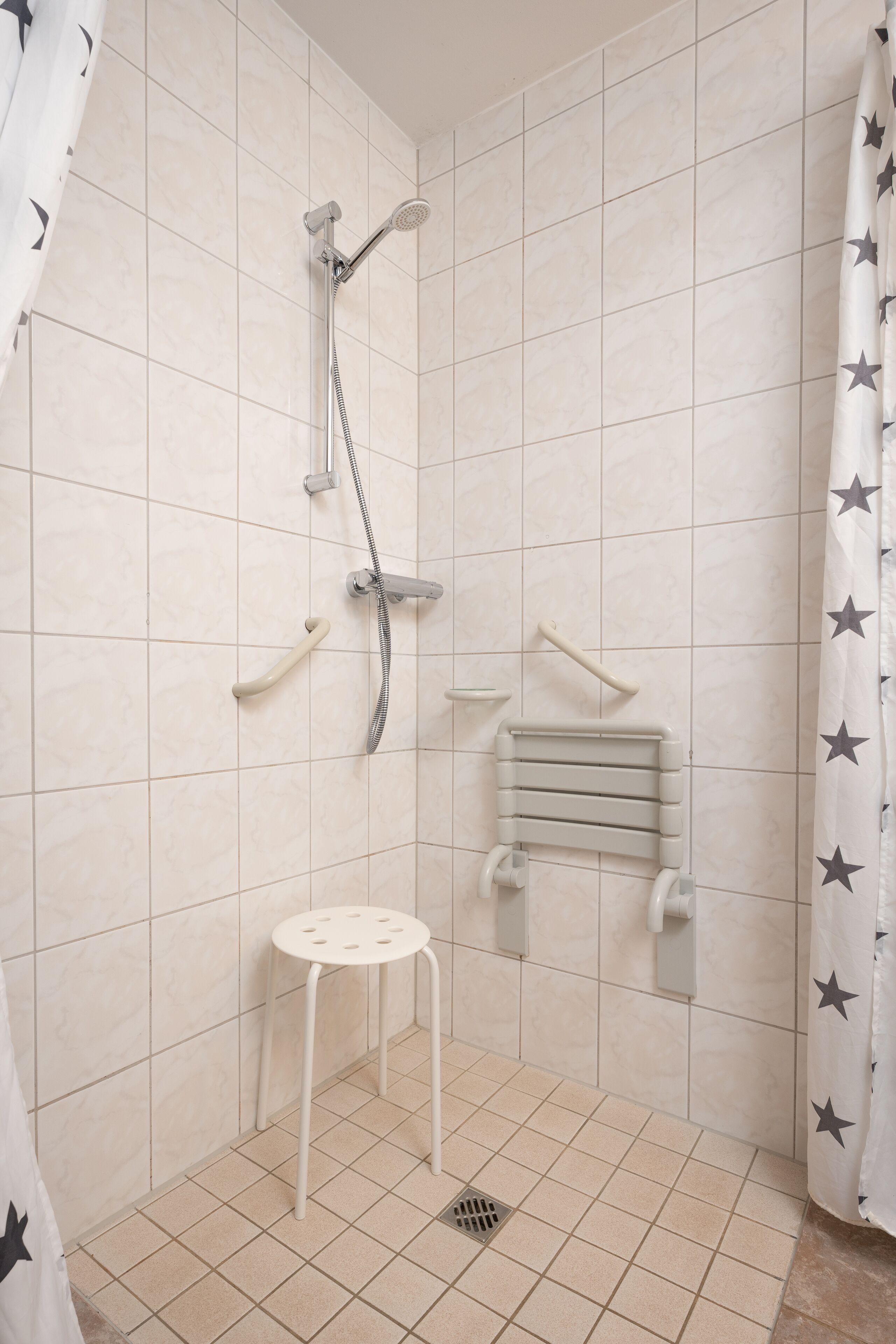 Un espacio de ducha pequeño con azulejos, un cabezal de ducha manual, un taburete pequeño y un perchero para toallas.