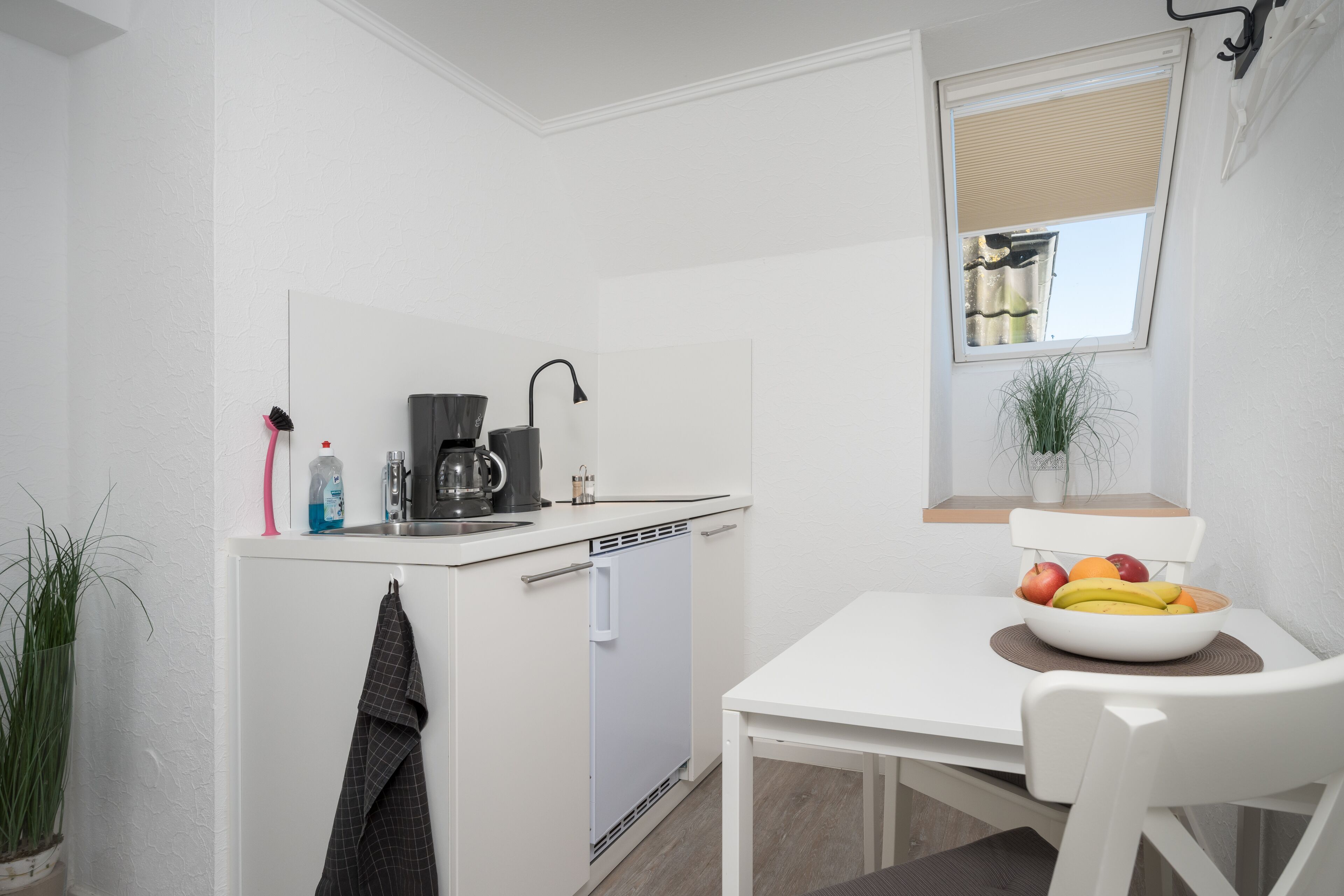 A compact kitchen with a white countertop, a coffee maker, and a bowl of fruit on a table.