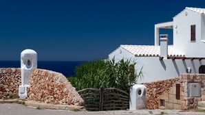 A white building with a stone wall and a gate, overlooking a clear blue sky and sea. - Apartment 4 In front of the sea (Urbanització Santo Tomás, Es Migjorn Gran)
