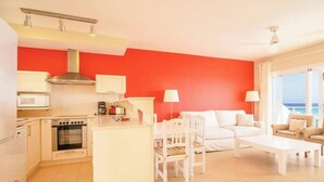 A compact living space with a kitchenette, a white sofa set, and a view of the sea through large windows. - Apartment 4 In front of the sea (Urbanització Santo Tomás, Es Migjorn Gran)