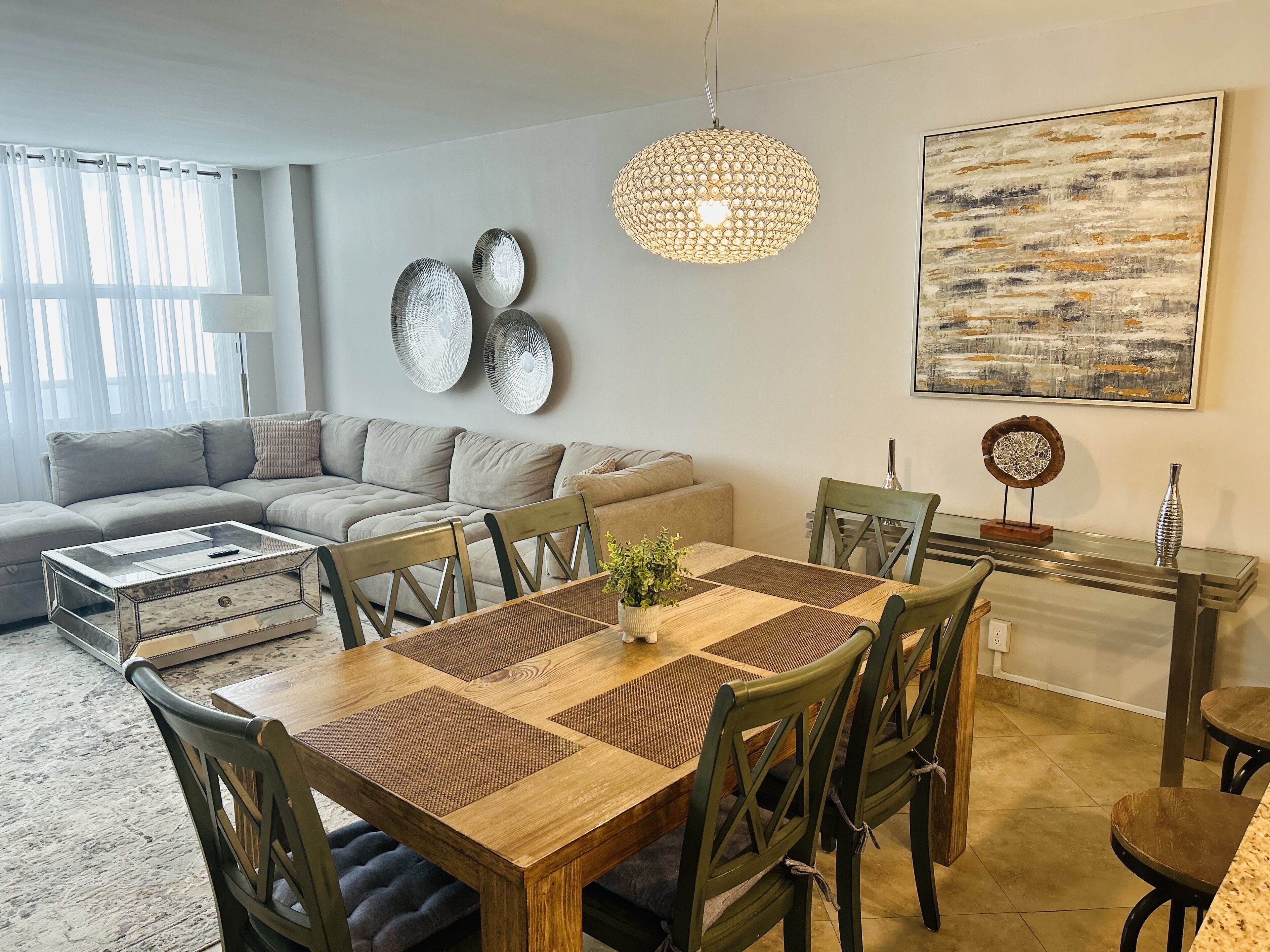 A modern living room with a wooden dining table, green chairs, a grey sofa, and a large abstract painting on the wall.