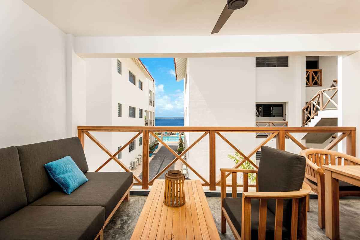 A balcony with a wooden table and chairs, a sofa with cushions, and a view of a narrow street and buildings.