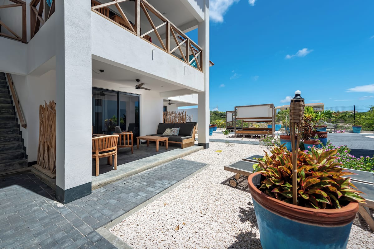 A modern two-story house with a balcony, outdoor seating area, and a large planter with plants.