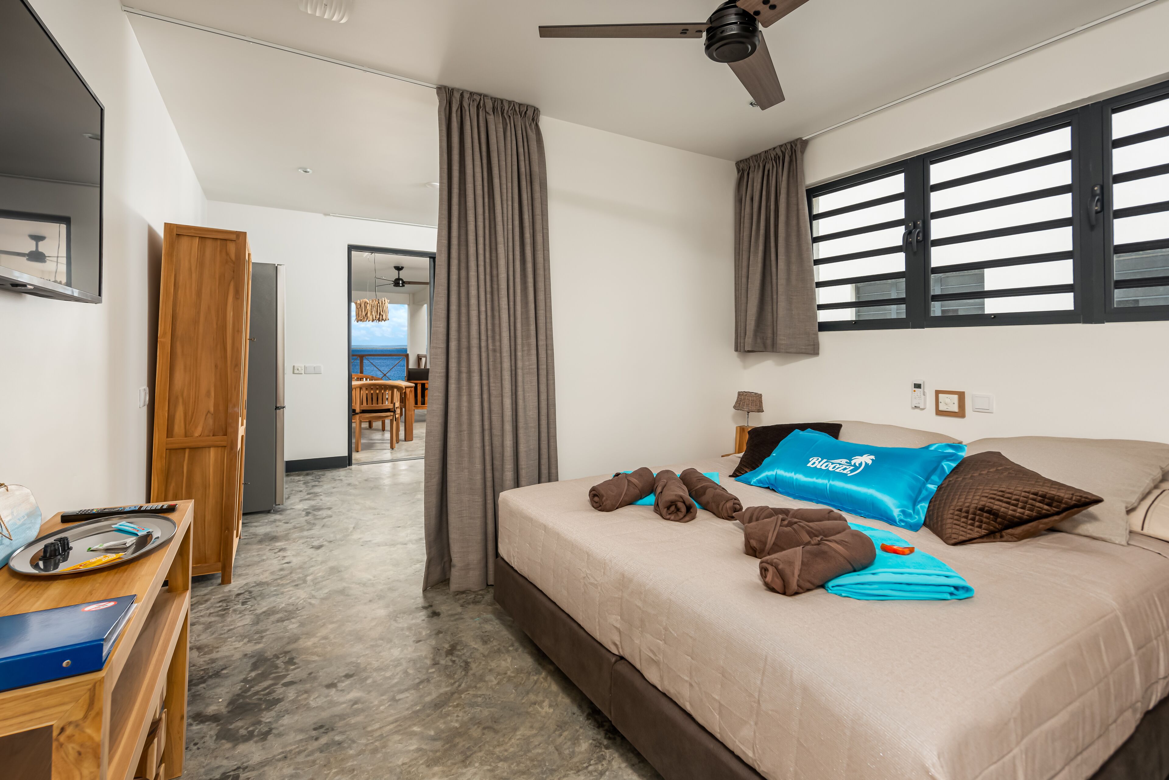 A hotel room with a bed, a desk, a ceiling fan, and a view of the sea through the window.