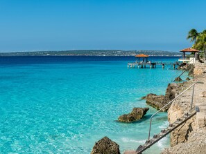 A coastal scene with clear turquoise waters, rocky shore, and a wooden pier with a gazebo. - Bloozz resort Bonaire (Kralendijk)