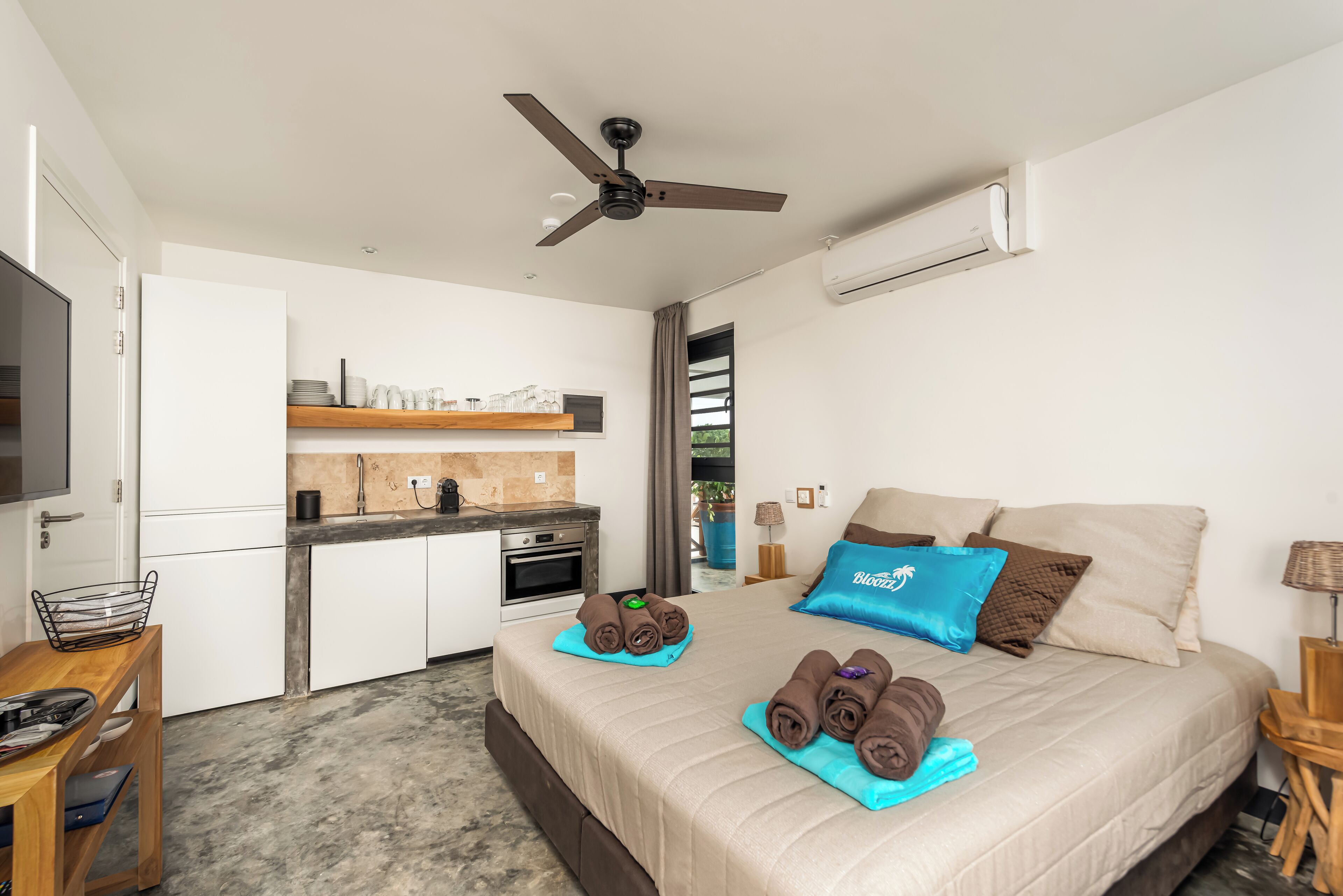 A modern hotel room with a bed, a small kitchenette, a ceiling fan, and an air conditioning unit.