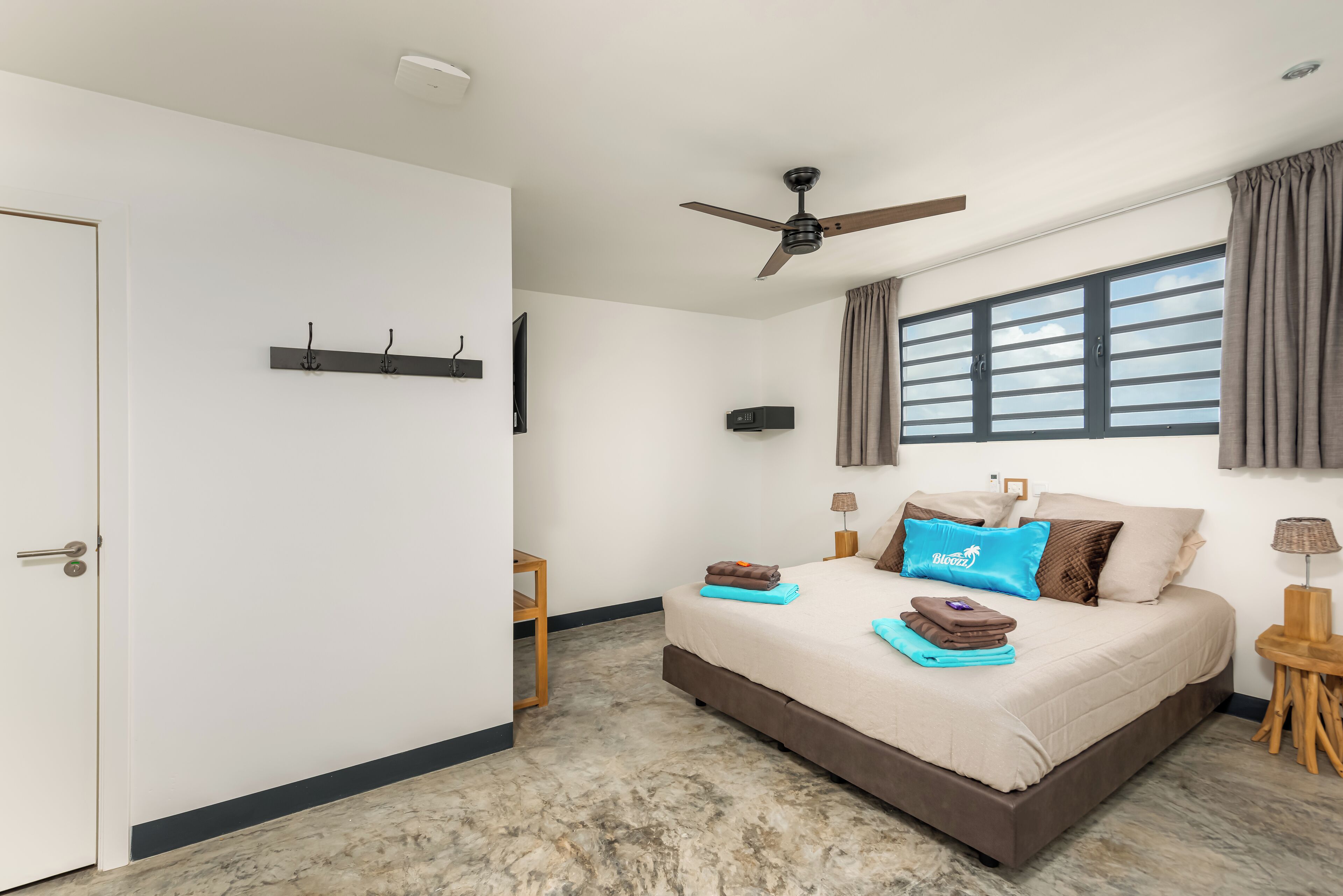Une chambre à coucher comprenant un lit, un ventilateur de plafond, une fenêtre avec des rideaux, un téléviseur fixé au mur et une porte.