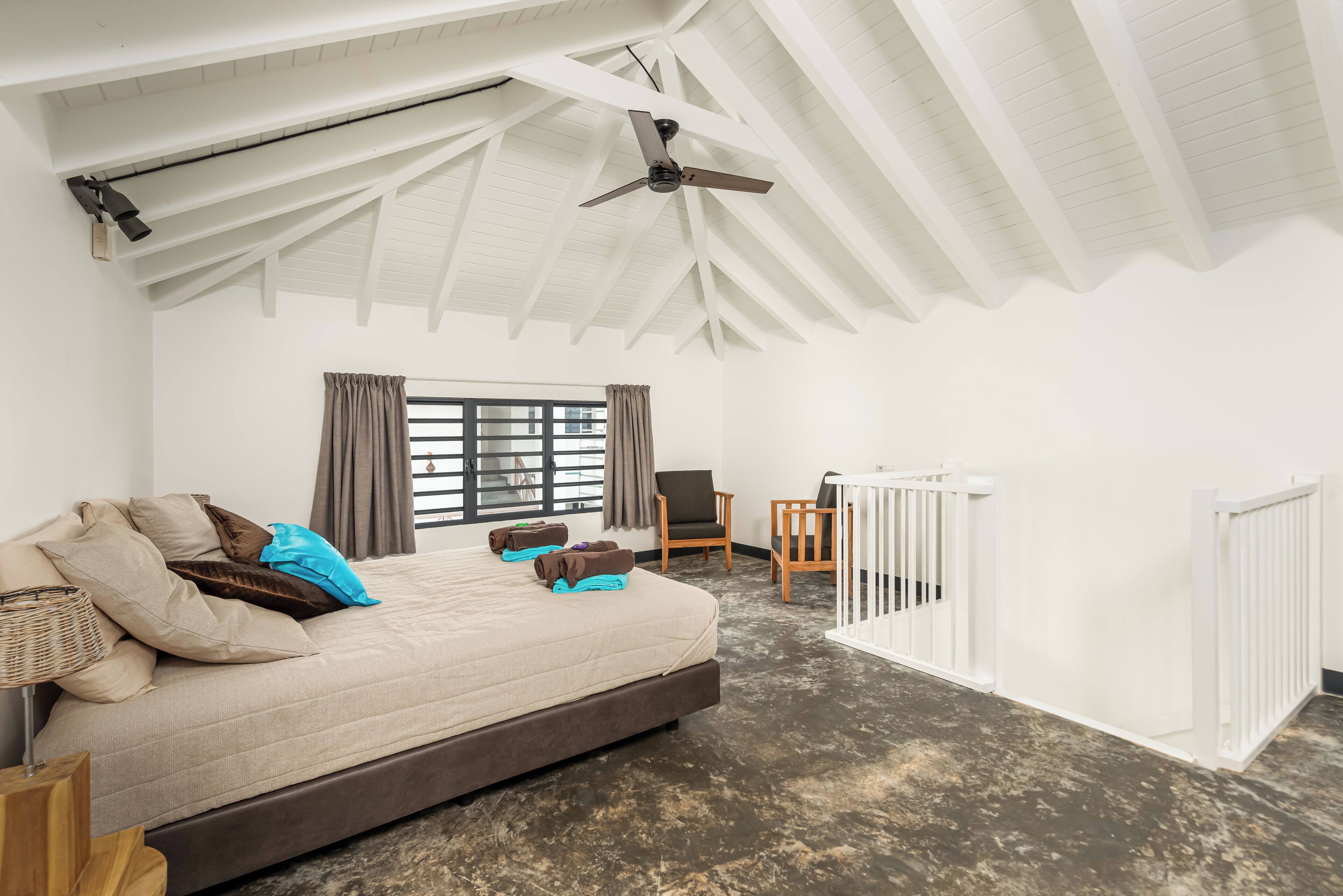 Un dormitorio amplio con una cama grande, un ventilador de techo, una ventana con cortinas, una silla y una barandilla blanca.
