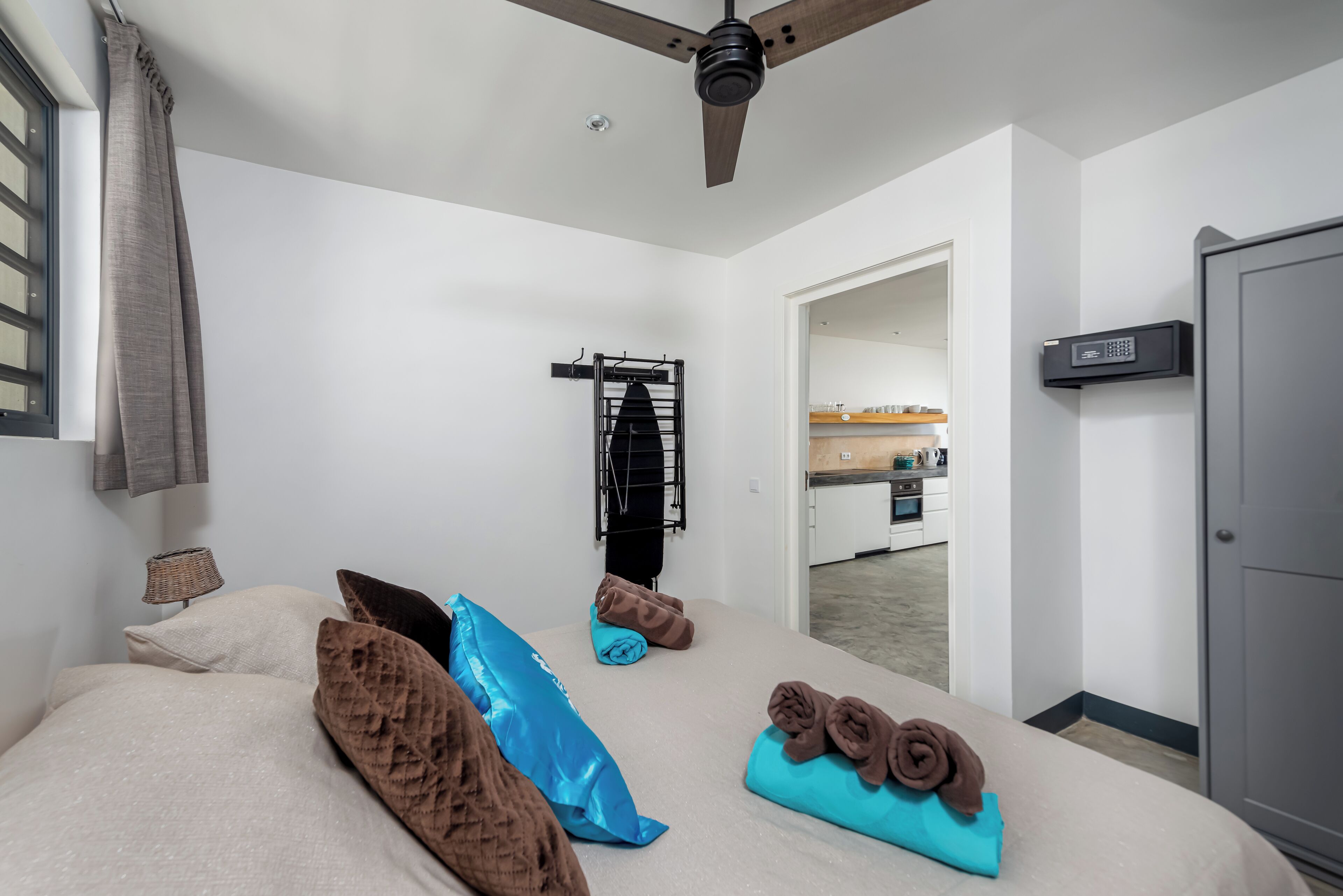 A bedroom with a bed, pillows, a ceiling fan, a towel rack, and a view into the kitchen.