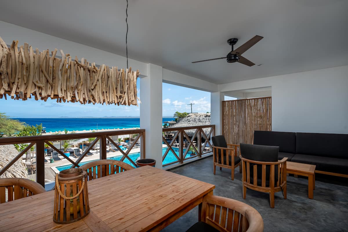 A wooden table and chairs on a balcony overlooking a pool and the ocean.