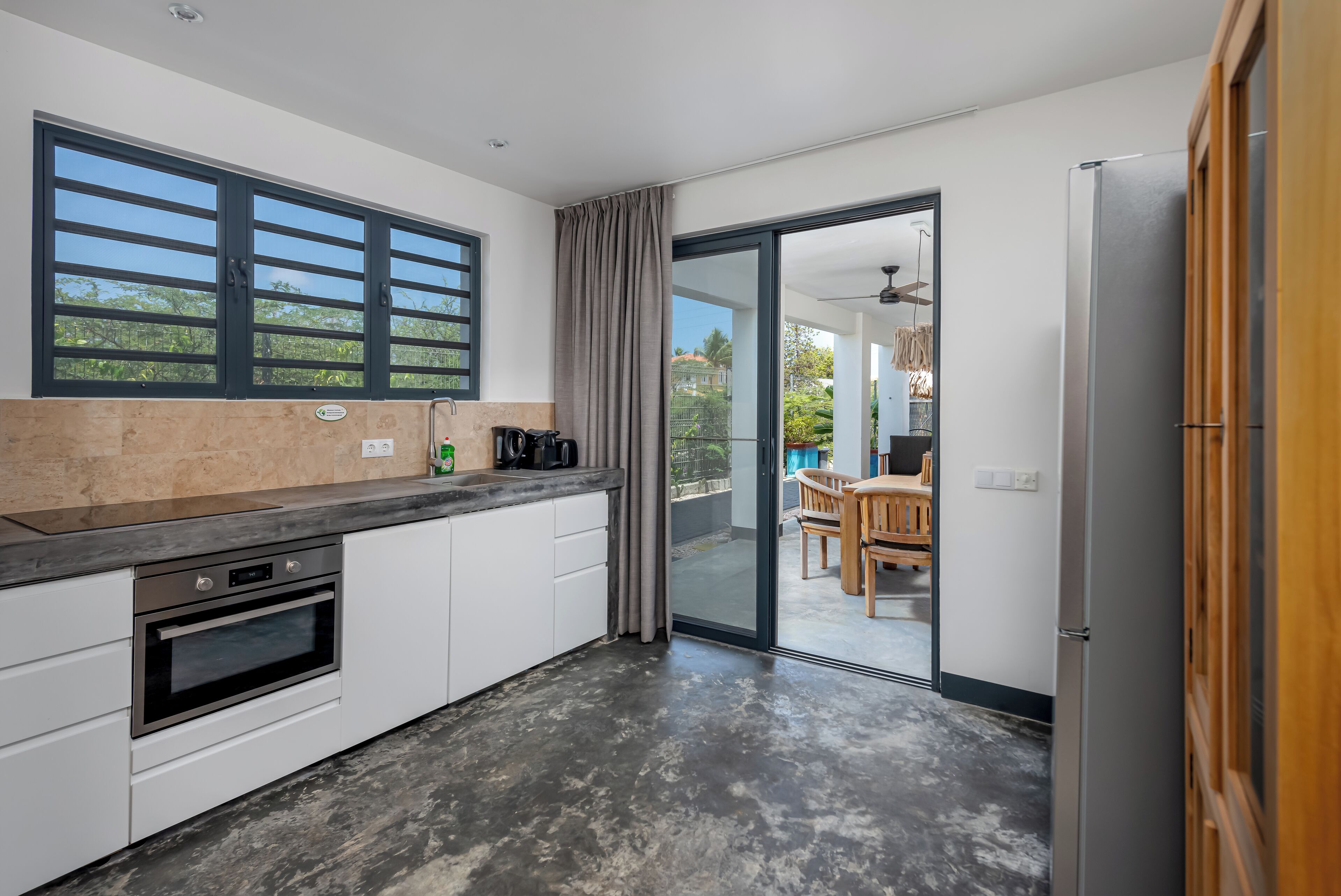 A modern kitchen with a built-in oven, a coffee maker, and a large window with black blinds.
