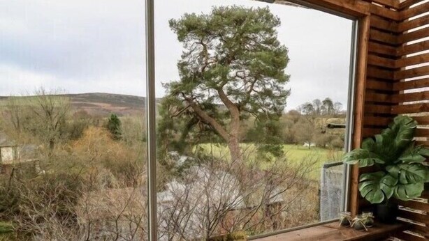 A room with a large window overlooking a river and trees. There is a wooden bench and a chair inside the room.