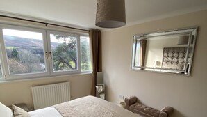 A bedroom with a large bed, a bedside table, a mirror, a window with curtains, and a view of the outdoors.