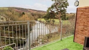 A patio with artificial grass, wicker furniture, a metal fence, and a view of a river and hills.