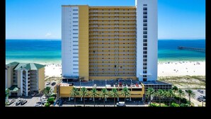Ein Hochhaus-Hotel mit gelb-weißer Fassade, Blick auf den Strand und ein Parkplatz mit Palmen.