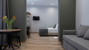 Une chambre moderne avec un canapé gris, une table ronde ornée d’un vase de fleurs, un lit recouvert d’une couverture grise et un téléviseur fixé au mur.