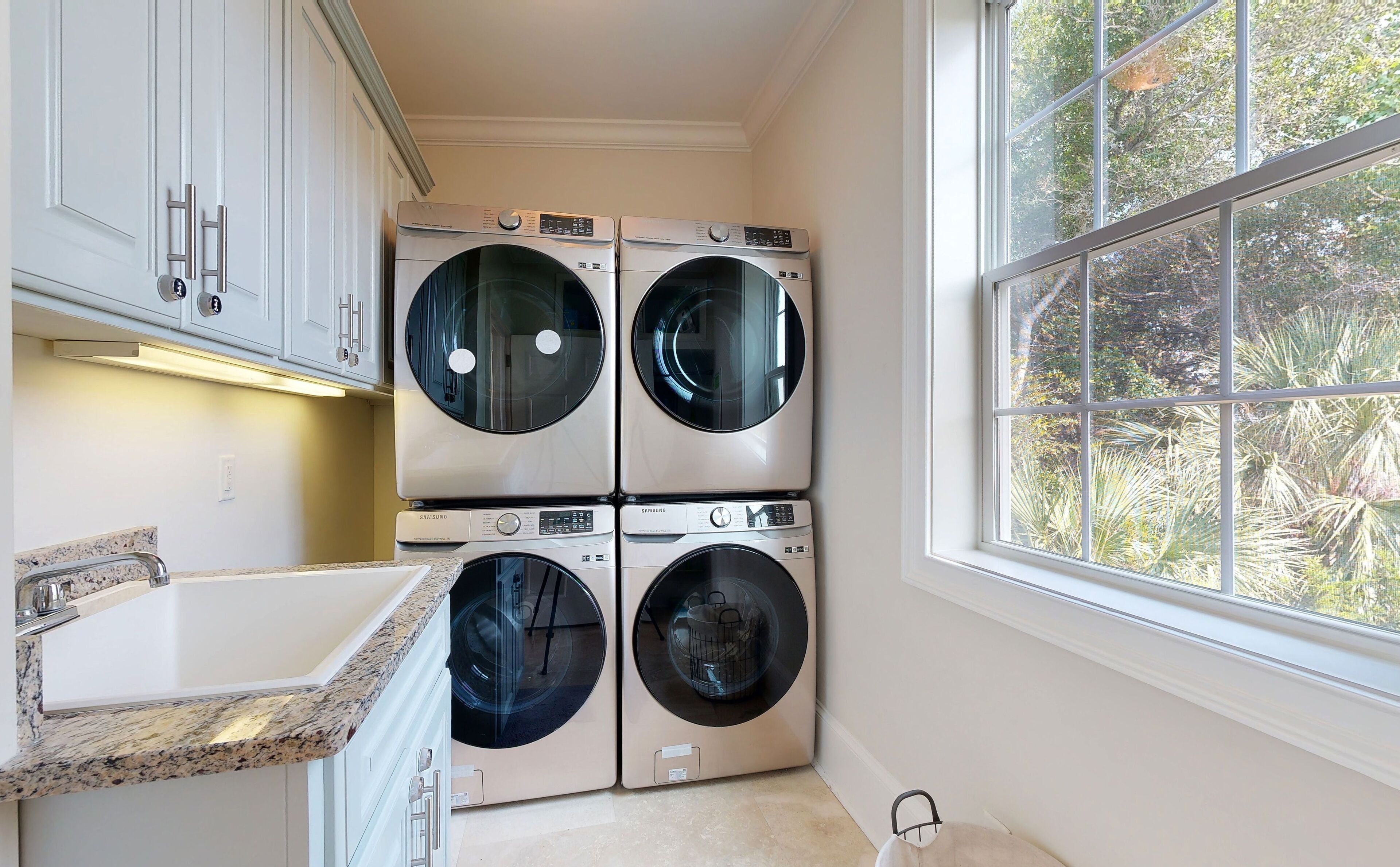 A laundry room with four Samsung washing machines, a granite countertop, and a window with a view of trees.