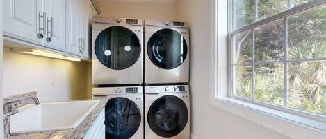 A laundry room with four Samsung washing machines, a granite countertop, and a window with a view of trees.