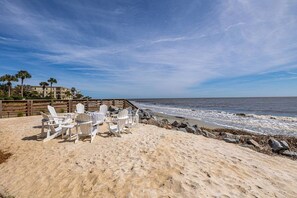 A beach with white Adirondack chairs, a wooden fence, and a building in the background.