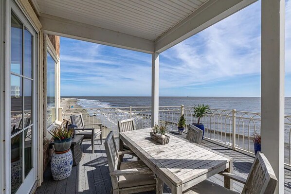 A covered outdoor area with a wooden table and chairs, potted plants, and a view of the ocean.
