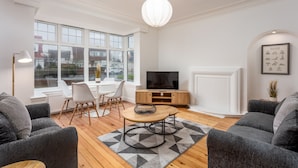 A modern living room with a TV, grey sofas, a wooden coffee table, and a large window with a view of buildings. - Wellington View - Donnini Apartments (Ayr)