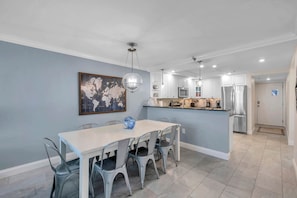 A dining area with a white table and metal chairs, a world map painting on the wall, and a kitchen in the background.