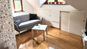 Ein modernes, helles Zimmer mit einem grauen Sofa, einem kleinen weißen Tisch und einem Fenster mit Blick auf Grünflächen.